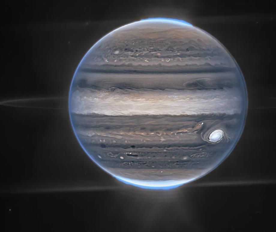 A composite image of Jupiter taken by the James Webb Space Telescope, showing the planet's rings and two of its moons, Amalthea and Adrastea. The blue glow around Jupiter's poles is the aurora.