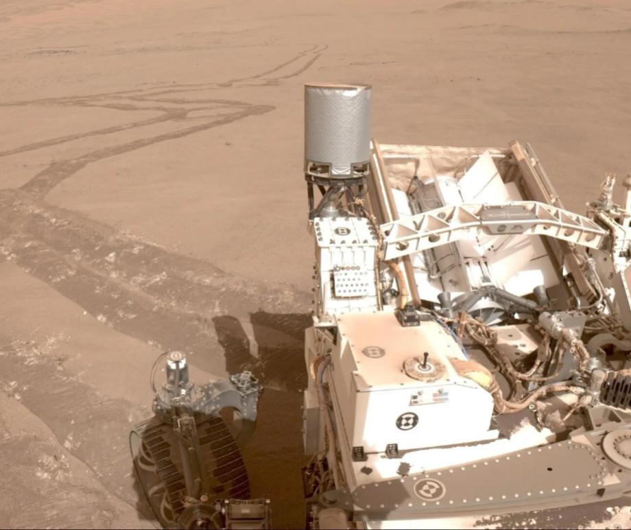 An image taken from one of Perseverance's cameras with the rover in the bottom right of the image and a long windy trail of tire tracks in the reddish brownish dirt of Mars behind it.